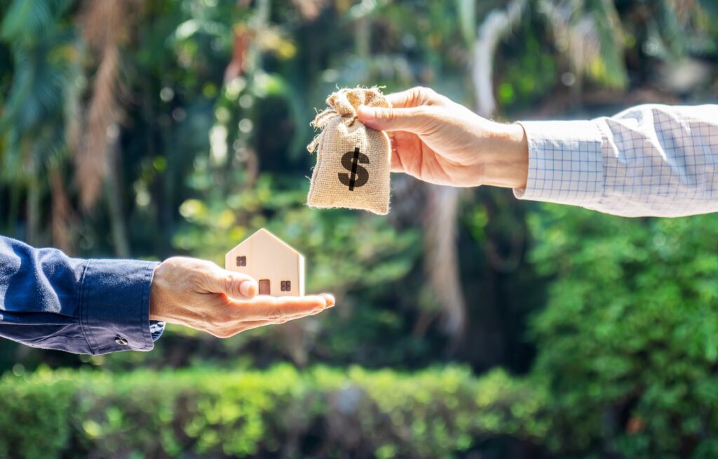 Small wooden house being held by someone with a blue shirt and a money bag being held by someone wearing a plad shirt